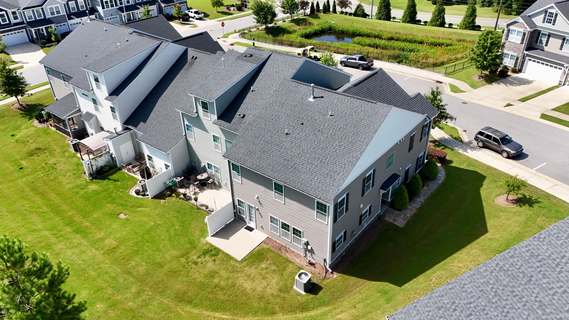 Aerial view of gray townhomes with dark gray roofs and green lawns; a road and pond are nearby.