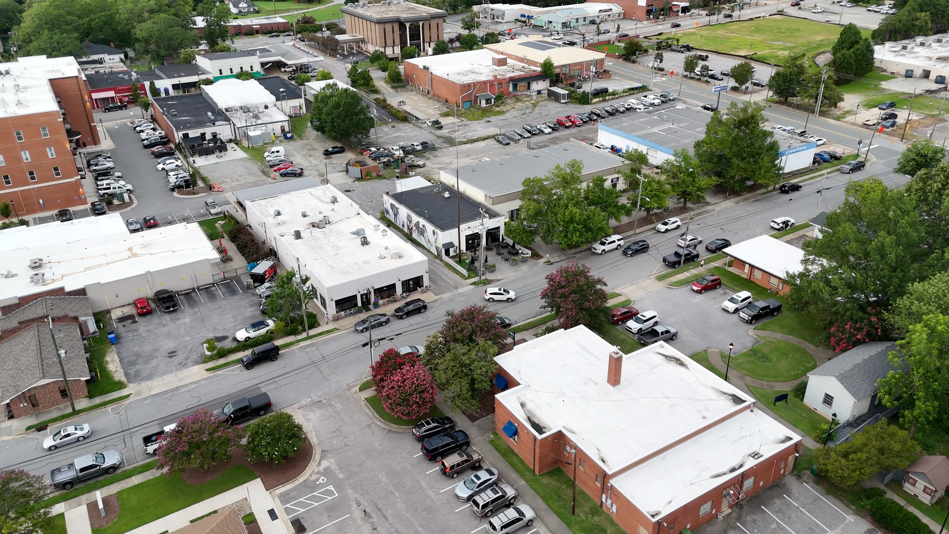 Aerial view of a town intersection with cars, buildings, and trees. Sunny day.