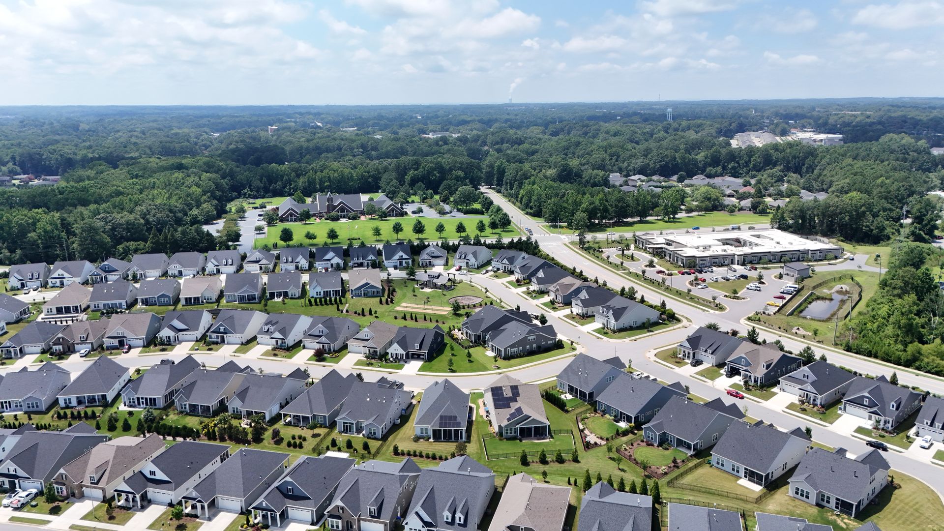 Aerial view of a suburban neighborhood with rows of houses, roads, and trees under a blue sky.