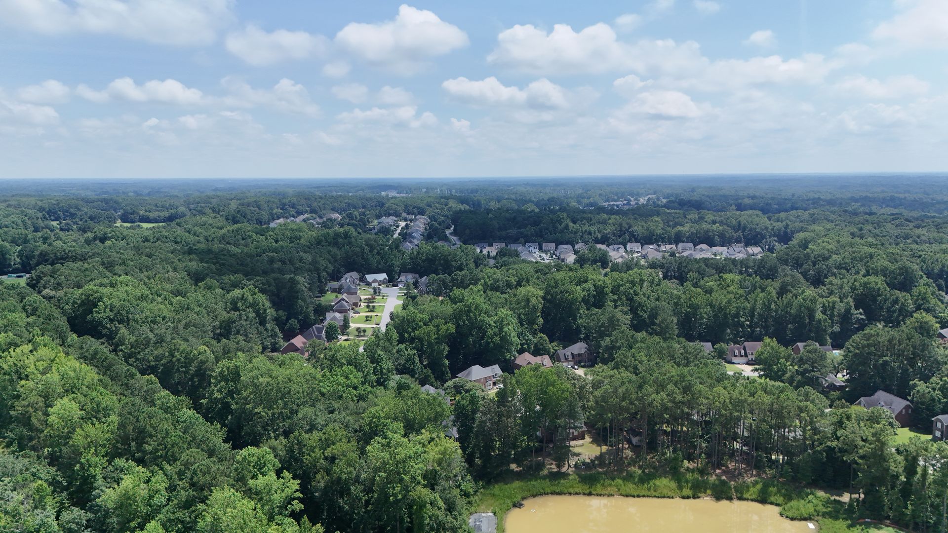 Overhead view of a green, tree-filled landscape with a neighborhood and pond visible under a blue sky.