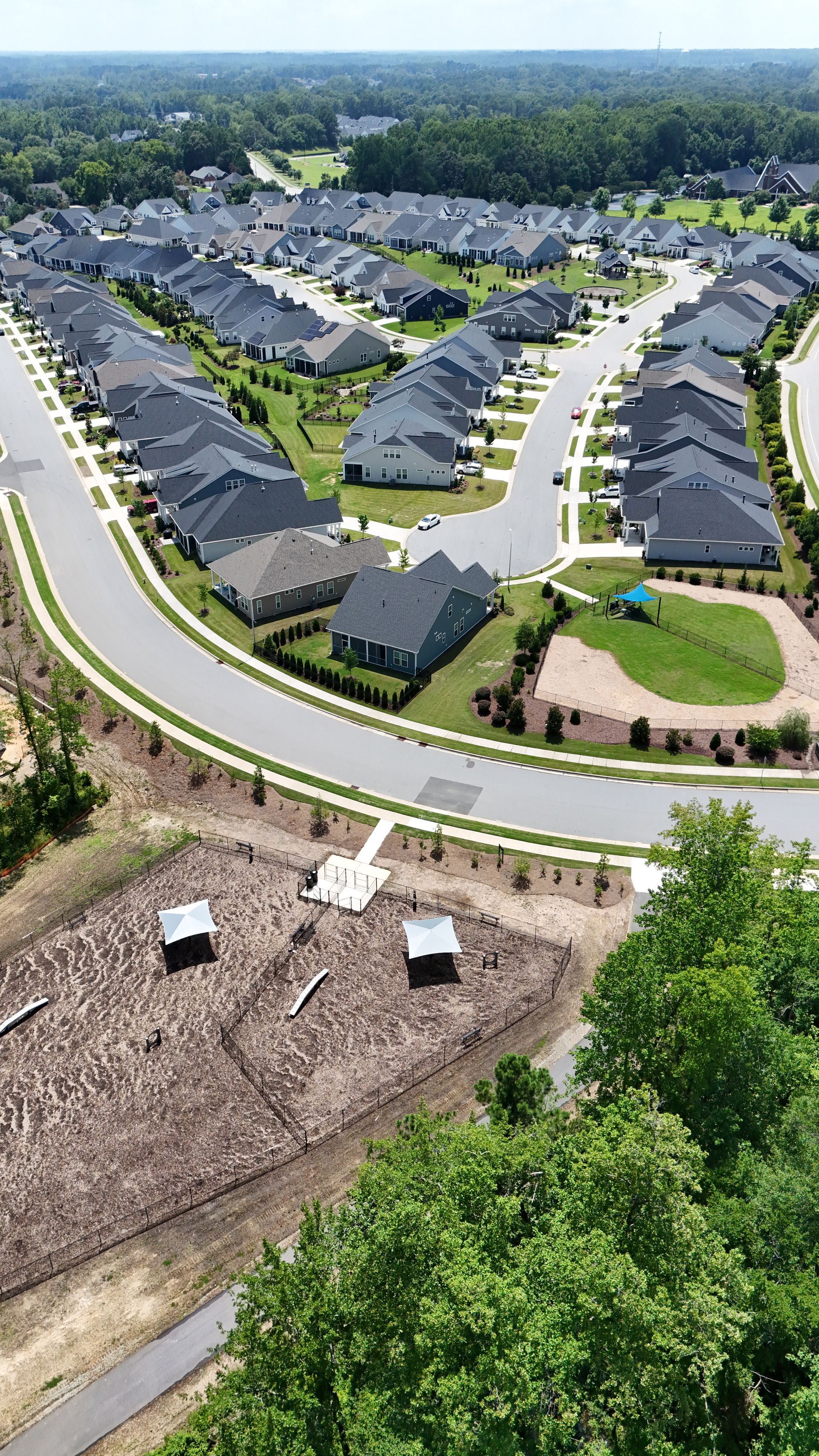 Aerial view of a suburban neighborhood with rows of houses and curved roads, near a wooded area with a small garden.