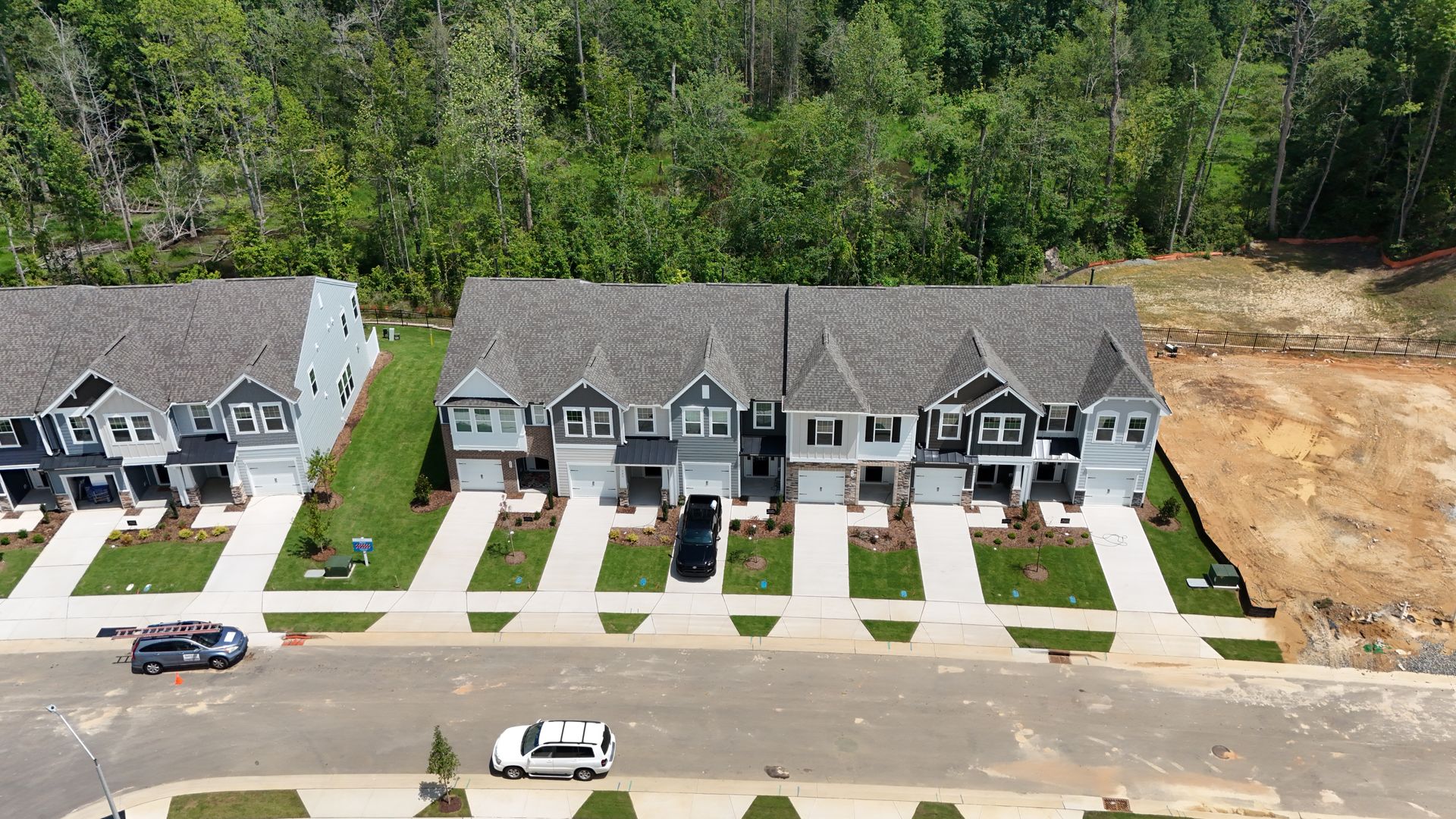 Row of townhouses in varying shades of gray with a forest in the background and parked cars on the street.