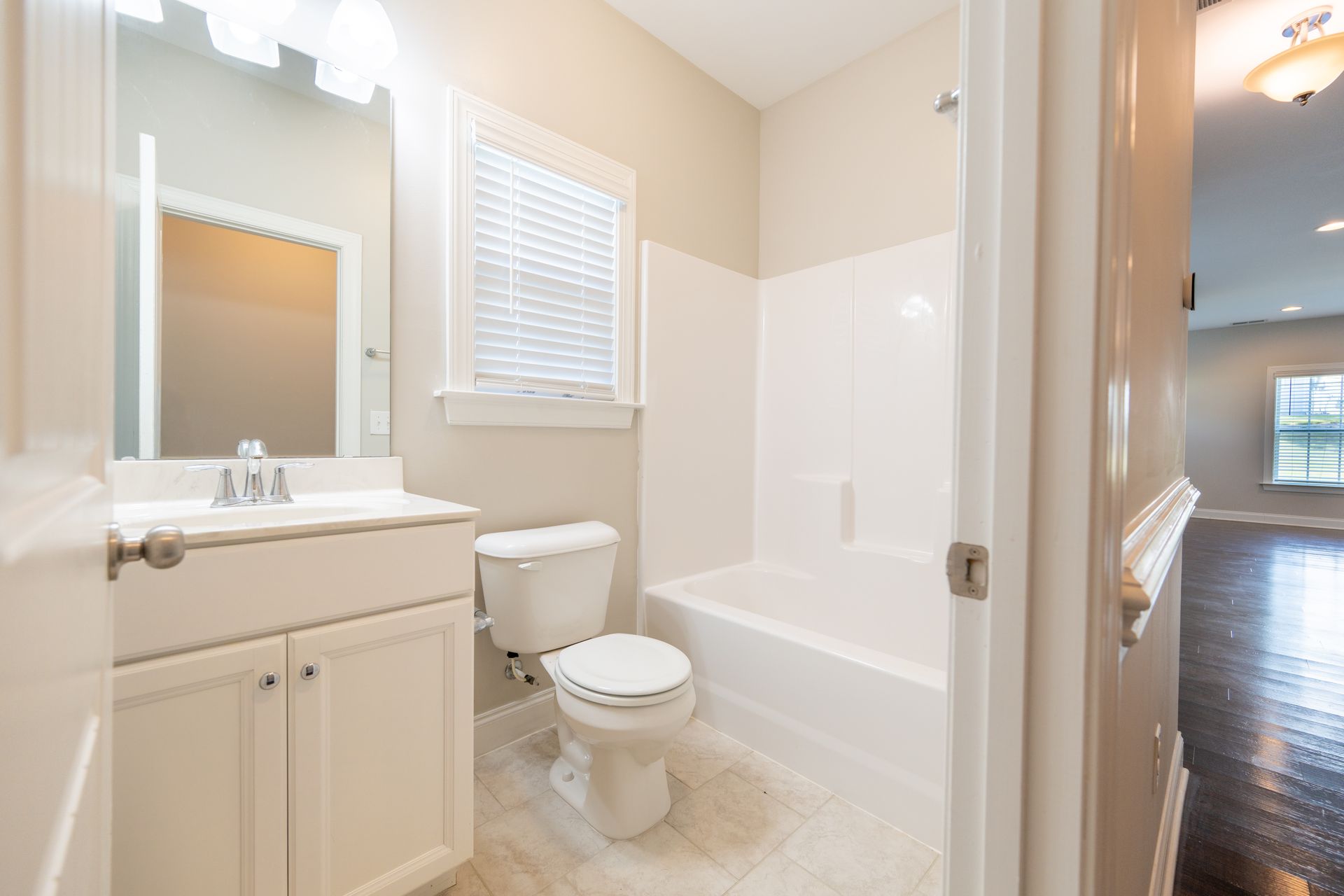 White bathroom with vanity, toilet, and bathtub; open door shows living room.