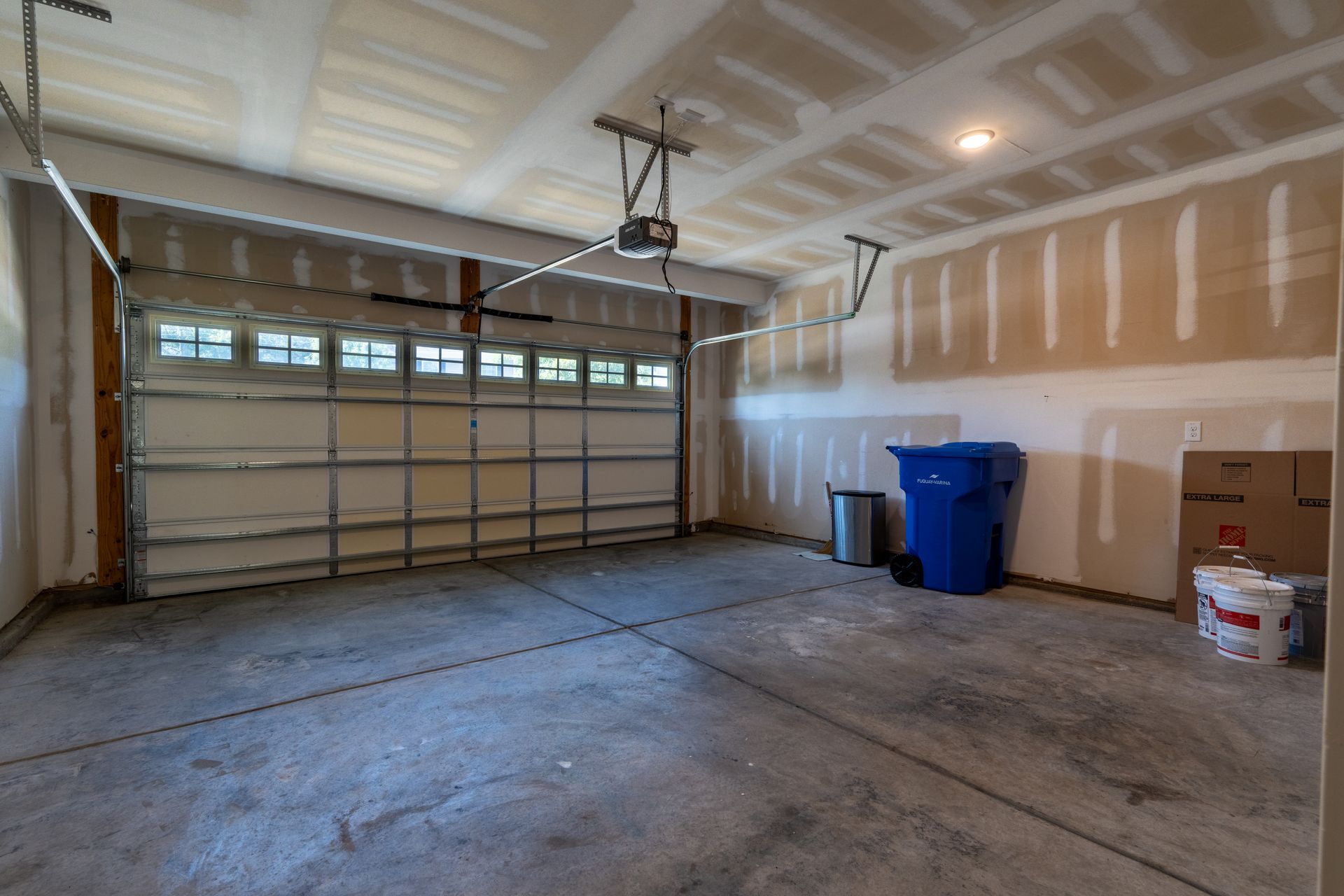 Empty garage with a closed garage door, unfinished walls, and a blue trash can.