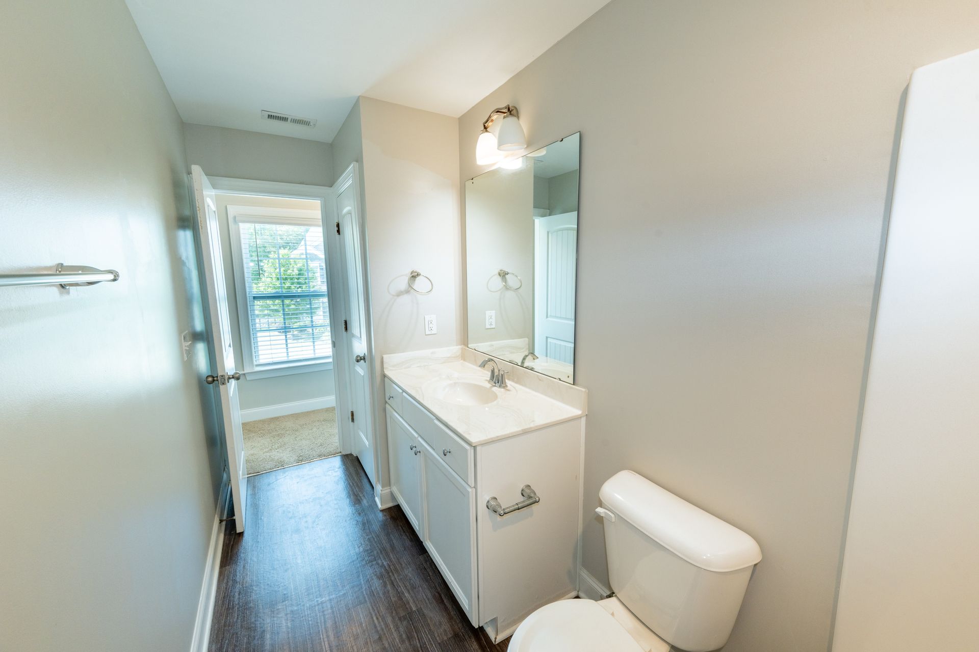 Narrow bathroom with white sink, toilet, and open door to exterior. Light gray walls, dark wood floor.