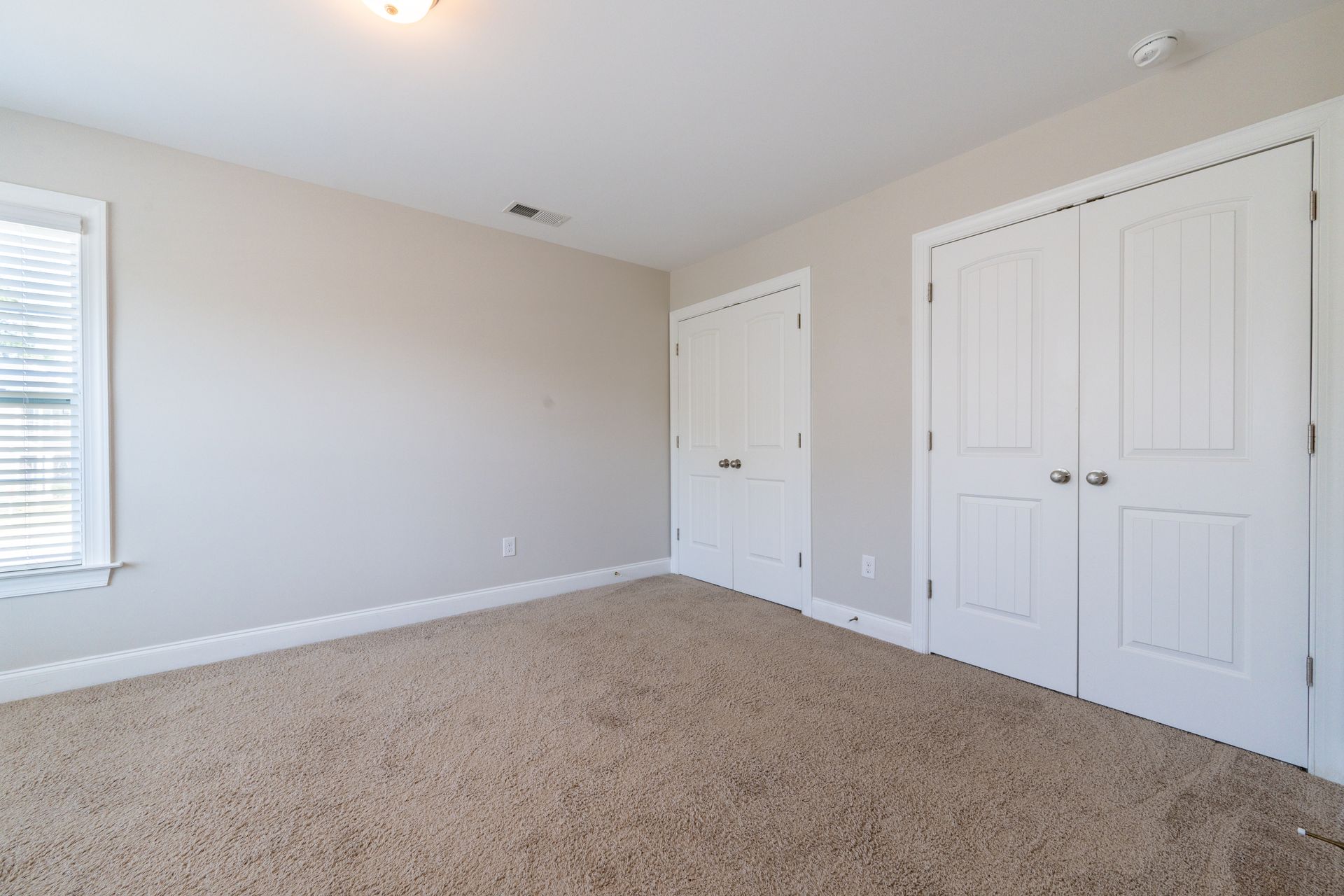 Empty bedroom with beige walls, tan carpet, white closet doors, and a window.