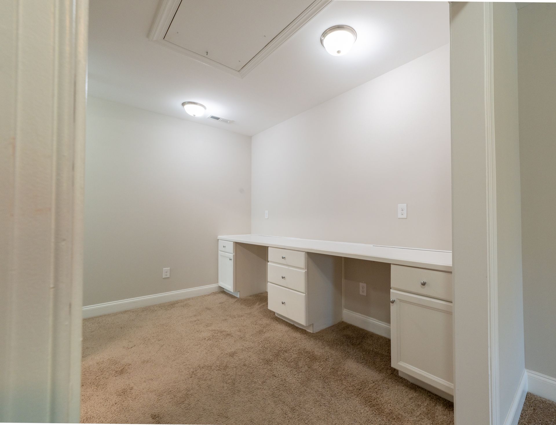 A small, empty office space with a built-in white desk and carpet flooring, lit by overhead lights.