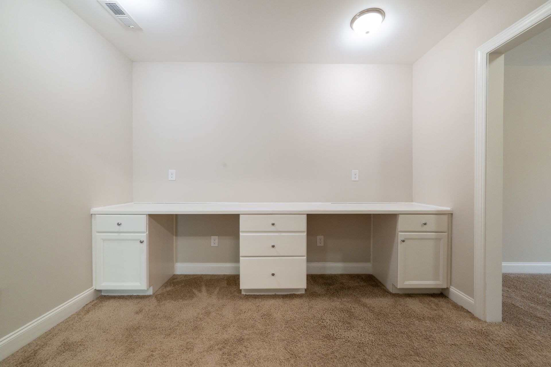 Built-in white desk with cabinets and drawers in a room with tan walls and carpet. Doorway on the right.