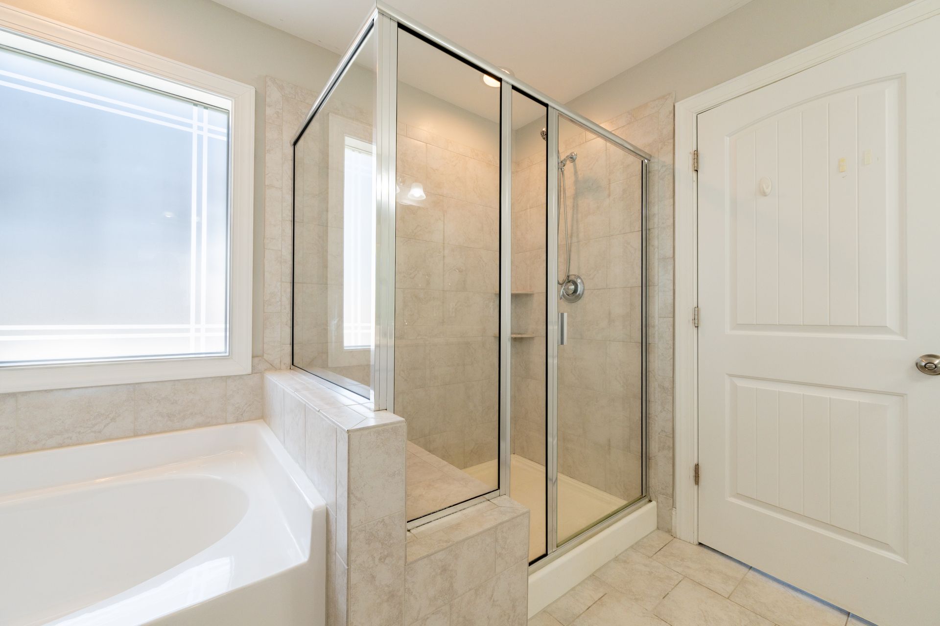 Bathroom with a clear glass shower, white door, and tub; light-colored walls and floor, window with blinds.