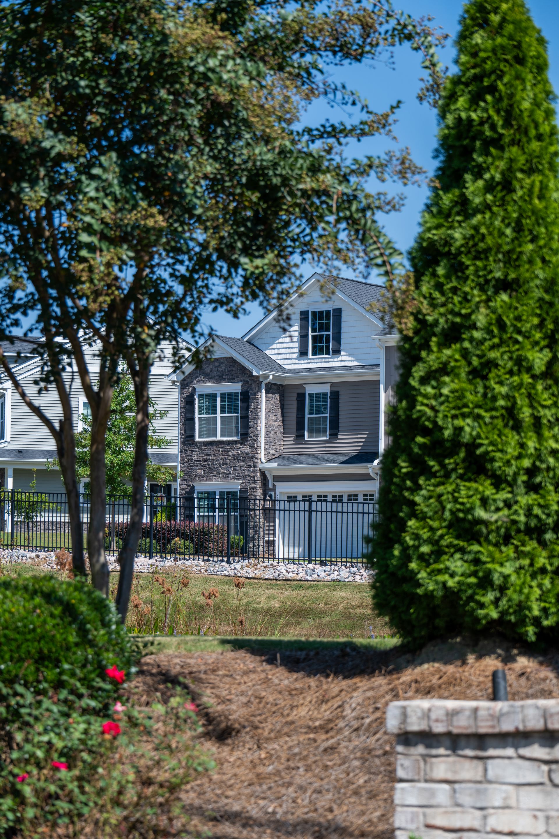 Two-story house with stone and siding facade, black shutters, behind a black fence and landscaping.