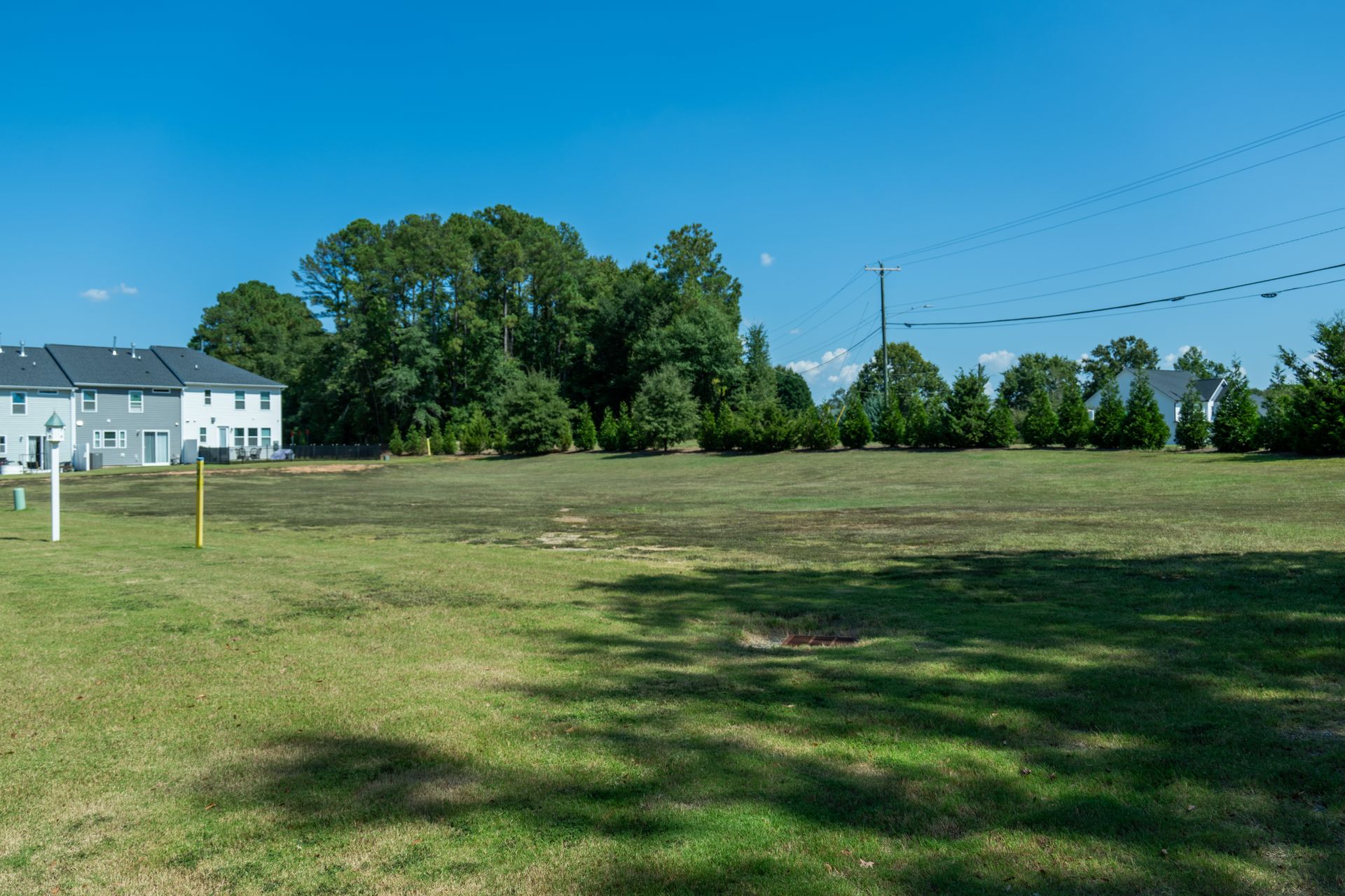 Grassy vacant lot with trees in the background and new townhouses on the left under a blue sky.