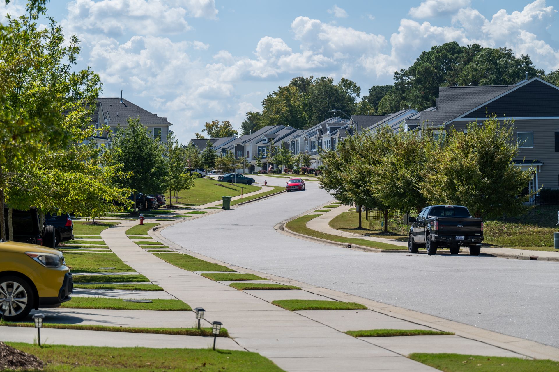 Suburban street with houses, trees, cars, and a cloudy sky.