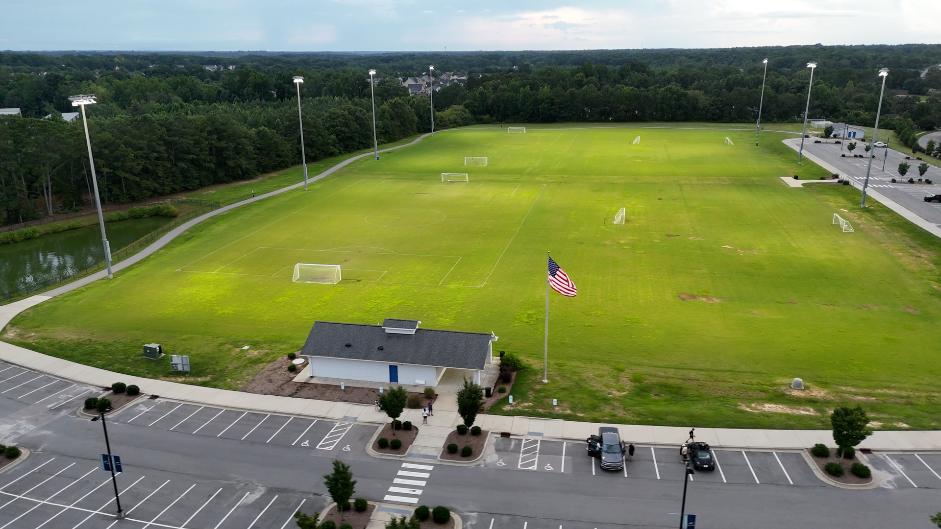 Soccer field under evening lights with a small building, American flag, and parking lot.