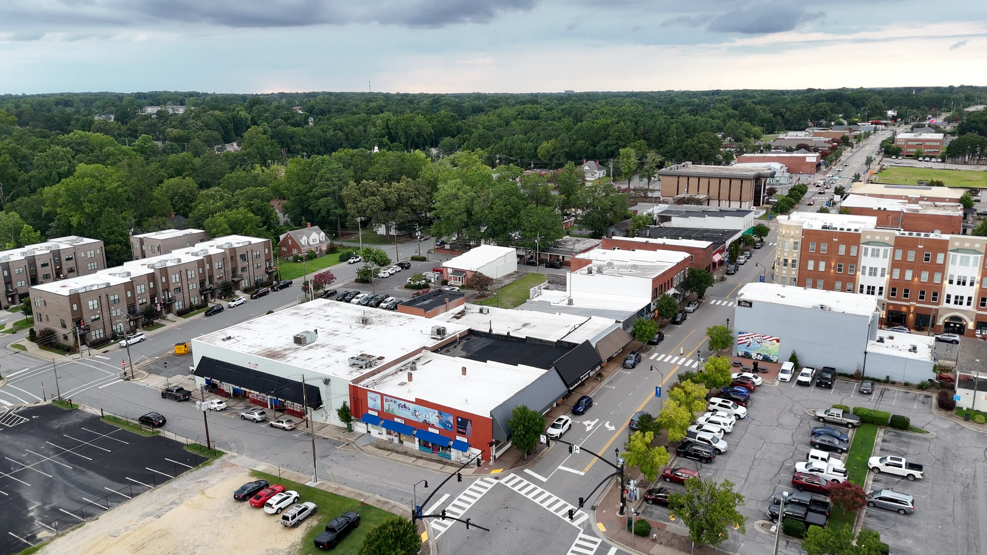 Aerial view of a small town street with shops, parked cars, and a forested area in the background.