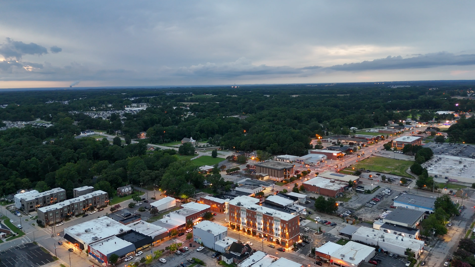 Aerial view of a town at dusk; buildings with lights in the foreground, dark forest in the background, overcast sky.