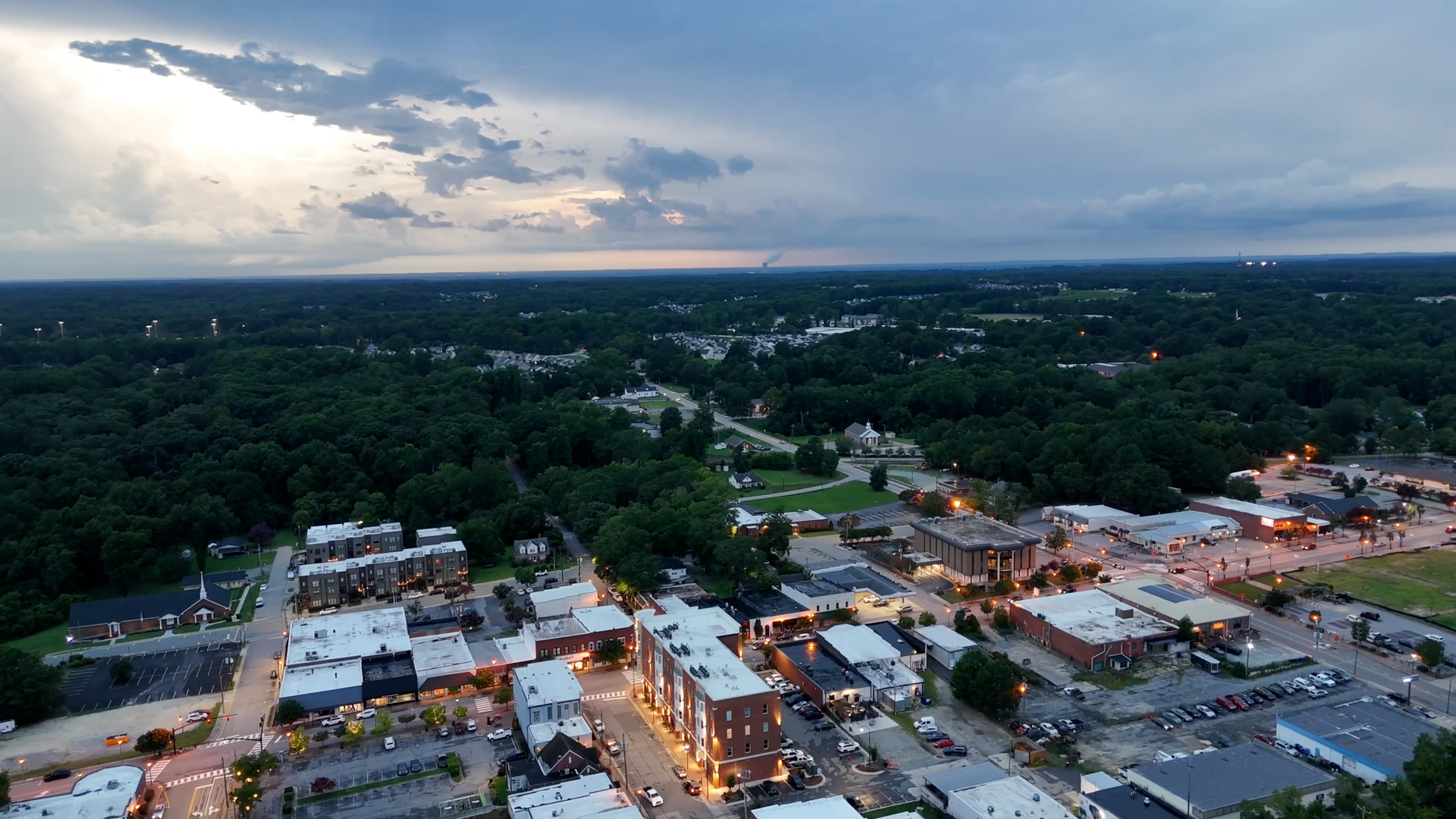 Aerial view of a town at dusk. Buildings with lights in front of a dark green forest under a cloudy sky.