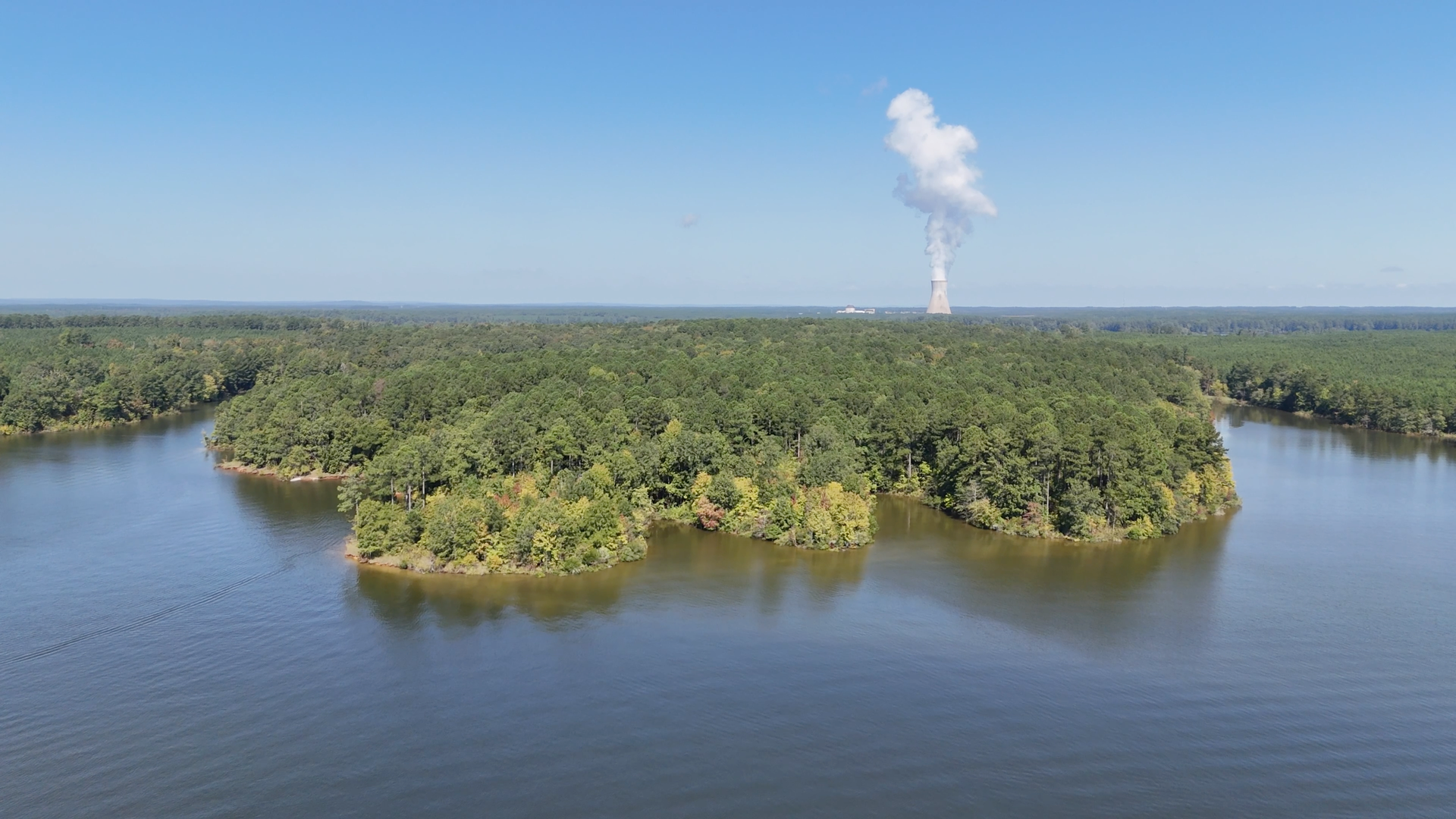 Lush green island in blue lake, with a tall smokestack emitting white smoke in the distance under a clear blue sky.