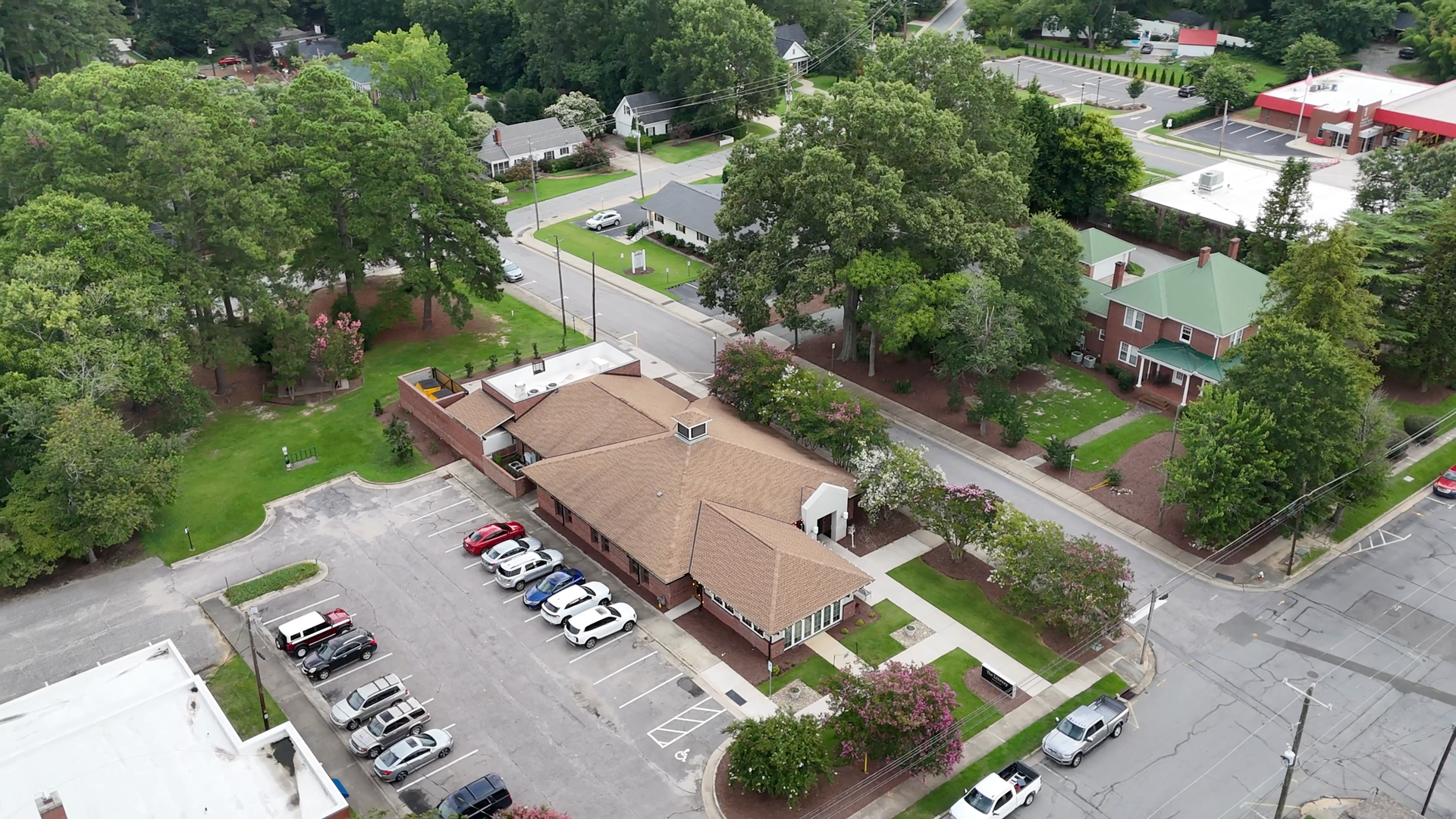 Aerial view of a building with brown roof and parking lot, surrounded by trees and streets.