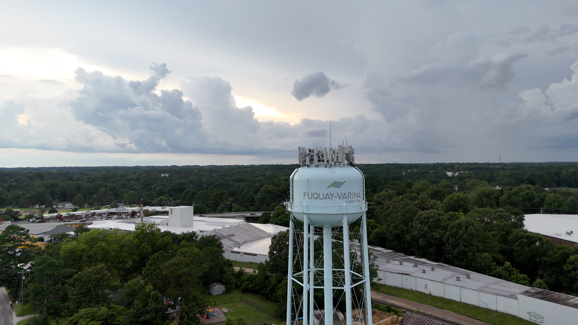 Water tower against a cloudy sky in a town. Trees surround the tower, with hints of buildings.