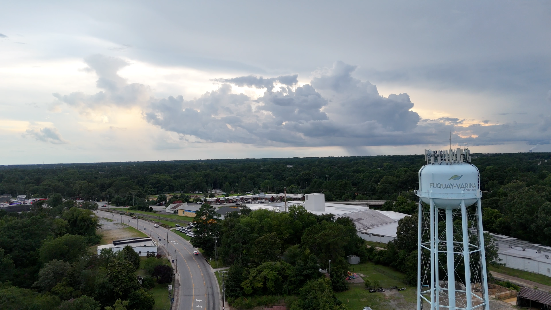 Water tower in front of cloudy sky over a town, trees, and road.