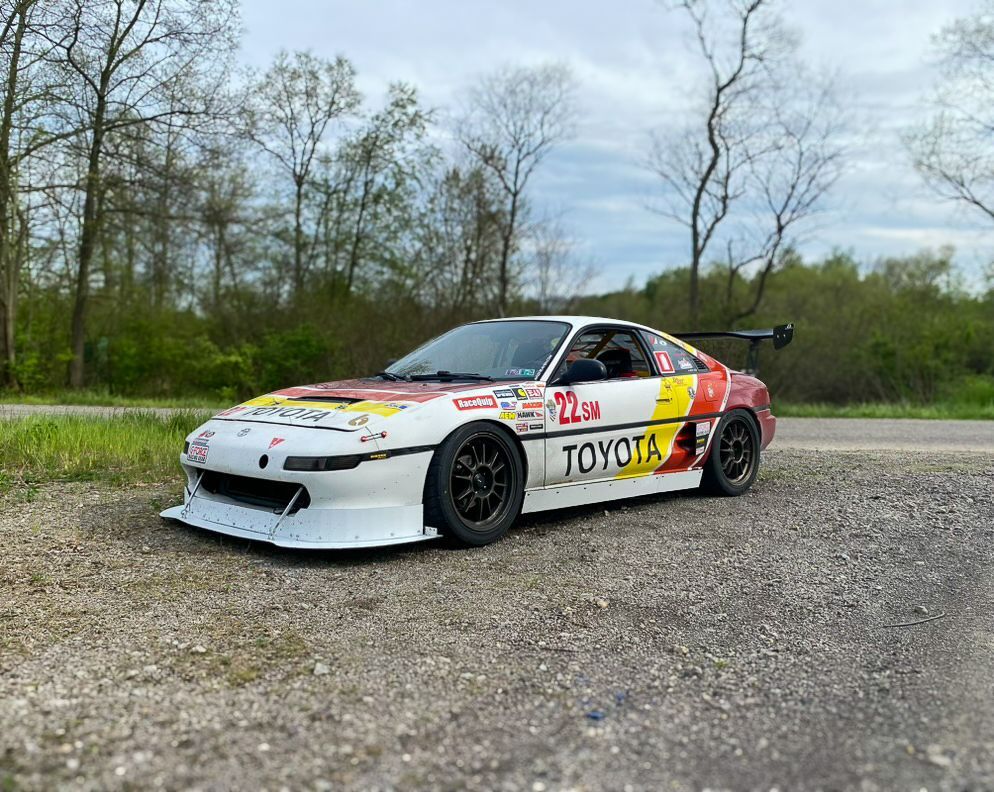 White and yellow Toyota MR2 race car on a gravel road. Black wheels, large spoiler, and racing livery.