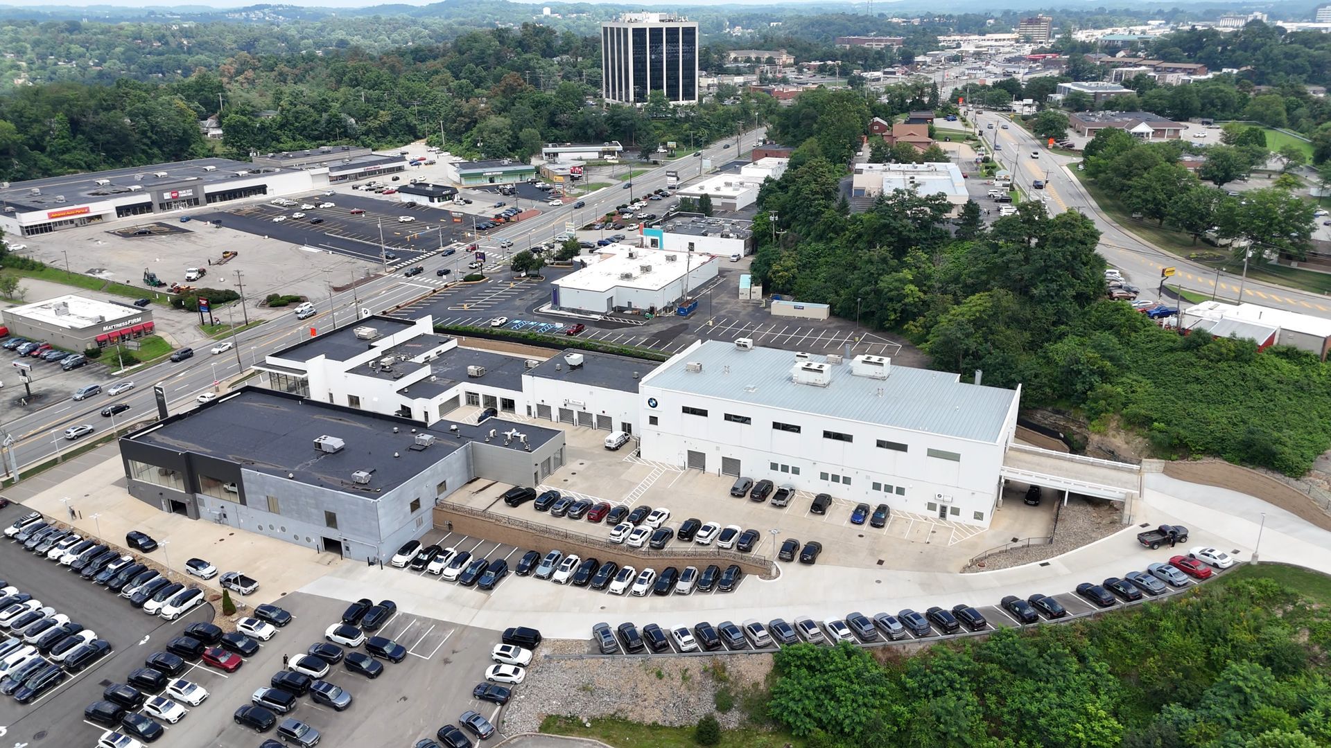Aerial view of commercial buildings and parking lots. Cars are parked throughout the lots. Trees are in the background.