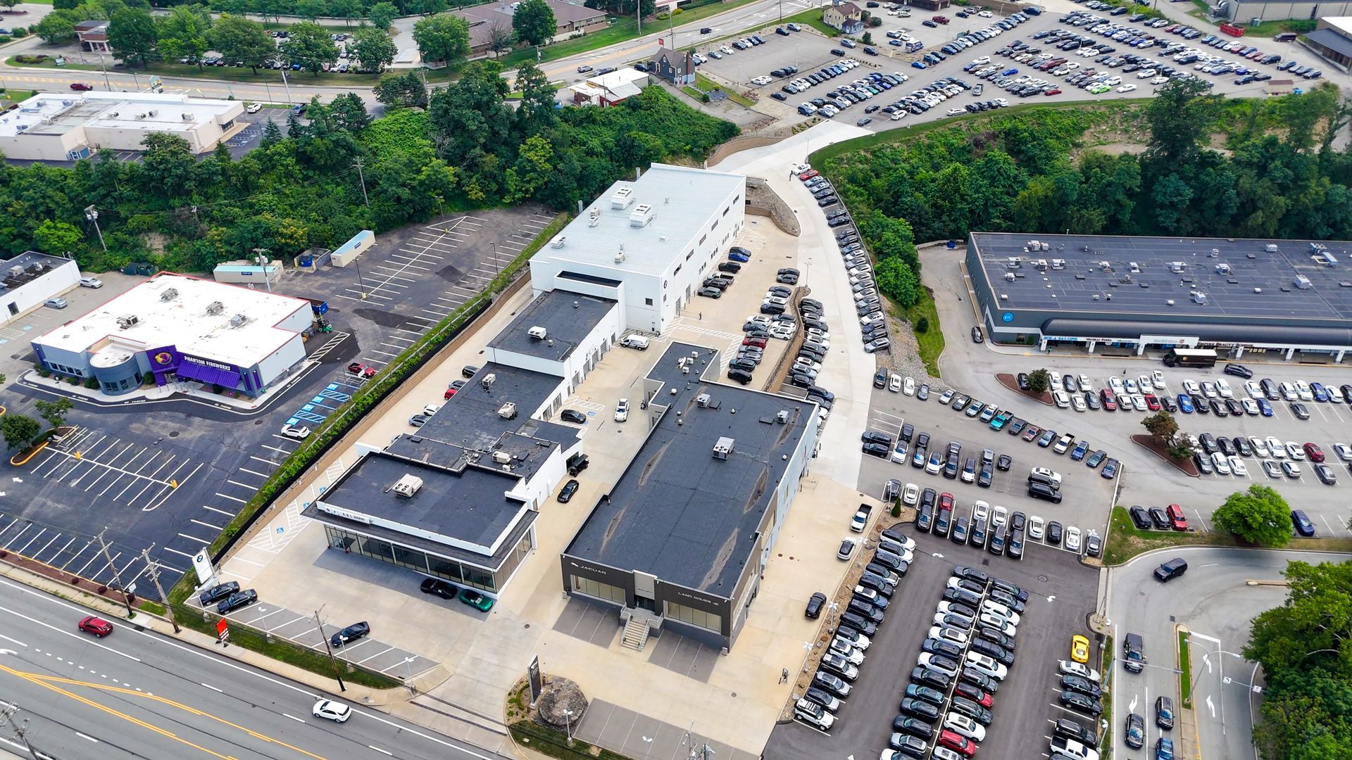 Aerial view of a commercial property with multiple buildings, parking lots, and vehicles. Green foliage and a road are visible.