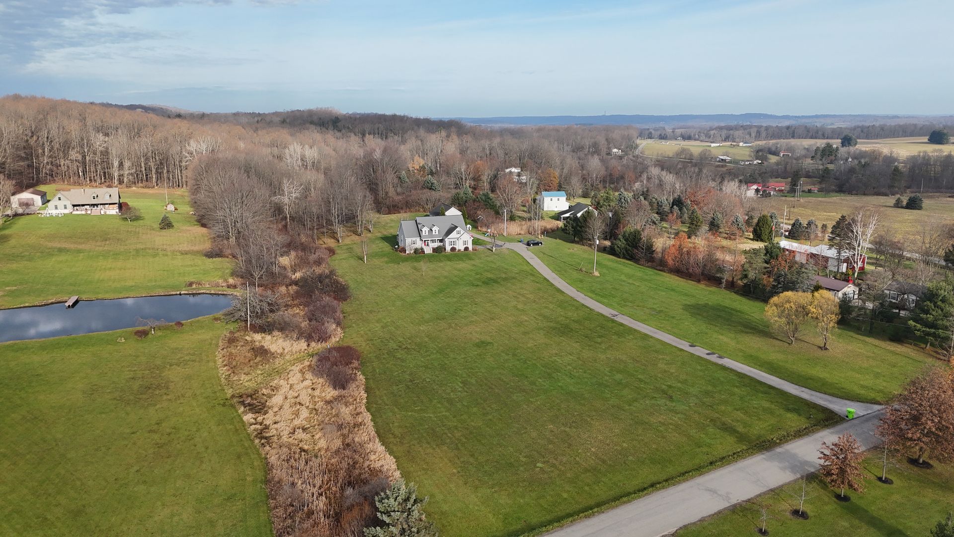 Aerial view of a house on a grassy hill near a pond and trees, under a clear blue sky.