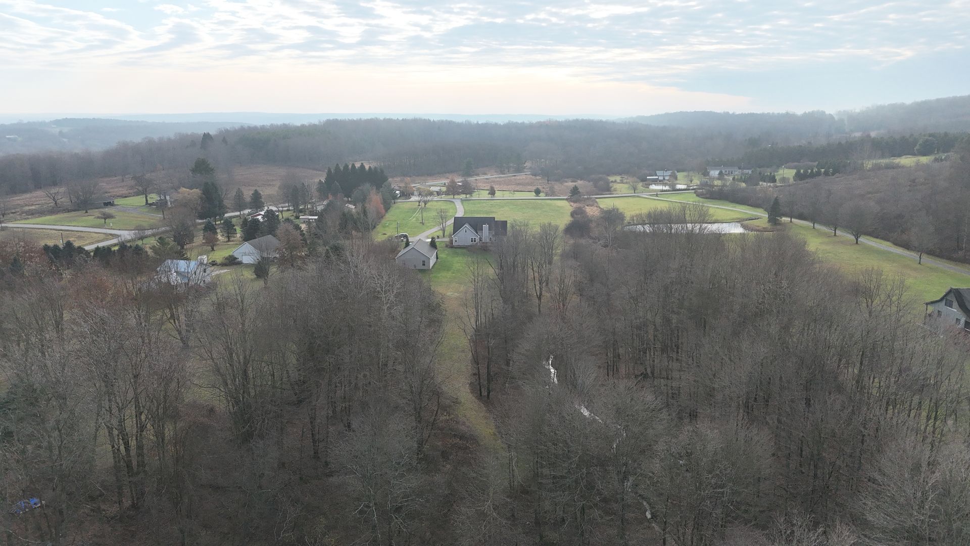 Hilly landscape with bare trees in foreground, houses dotting the distance, overcast sky.