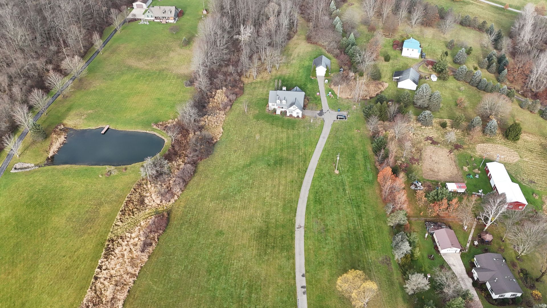 Aerial view: green hills with a pond, houses, and a long pathway in a rural setting.