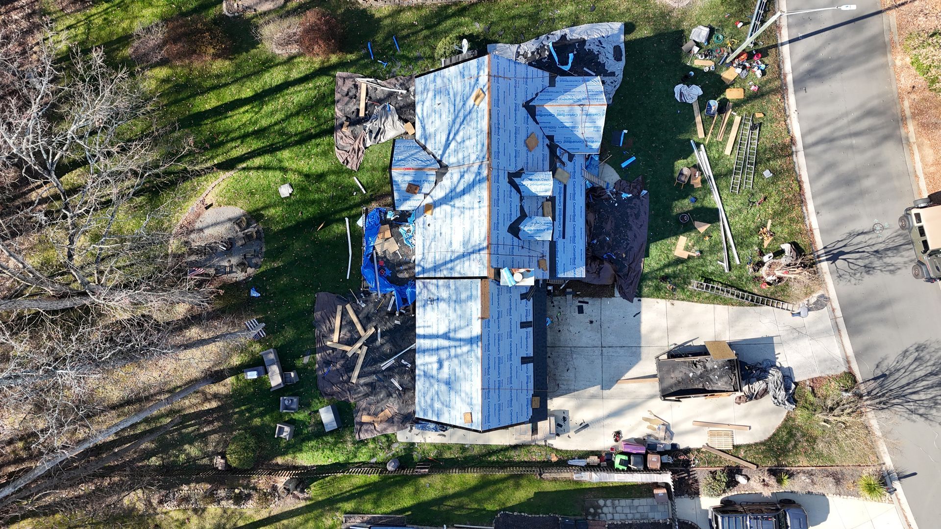 Aerial view of a house undergoing roof replacement, with scattered materials, surrounded by green grass and a road.