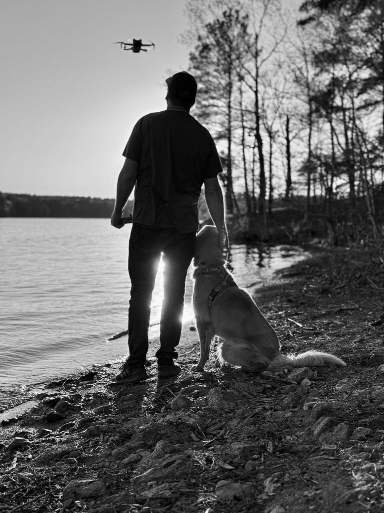 Man and dog at a lake watching a drone. Black and white. Sun sets behind them.