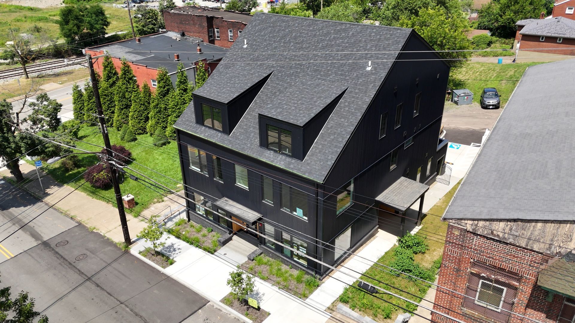 Modern, black three-story house with dormers, next to a brick building and greenery.