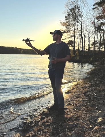 Man holding a drone near a lake at sunset.