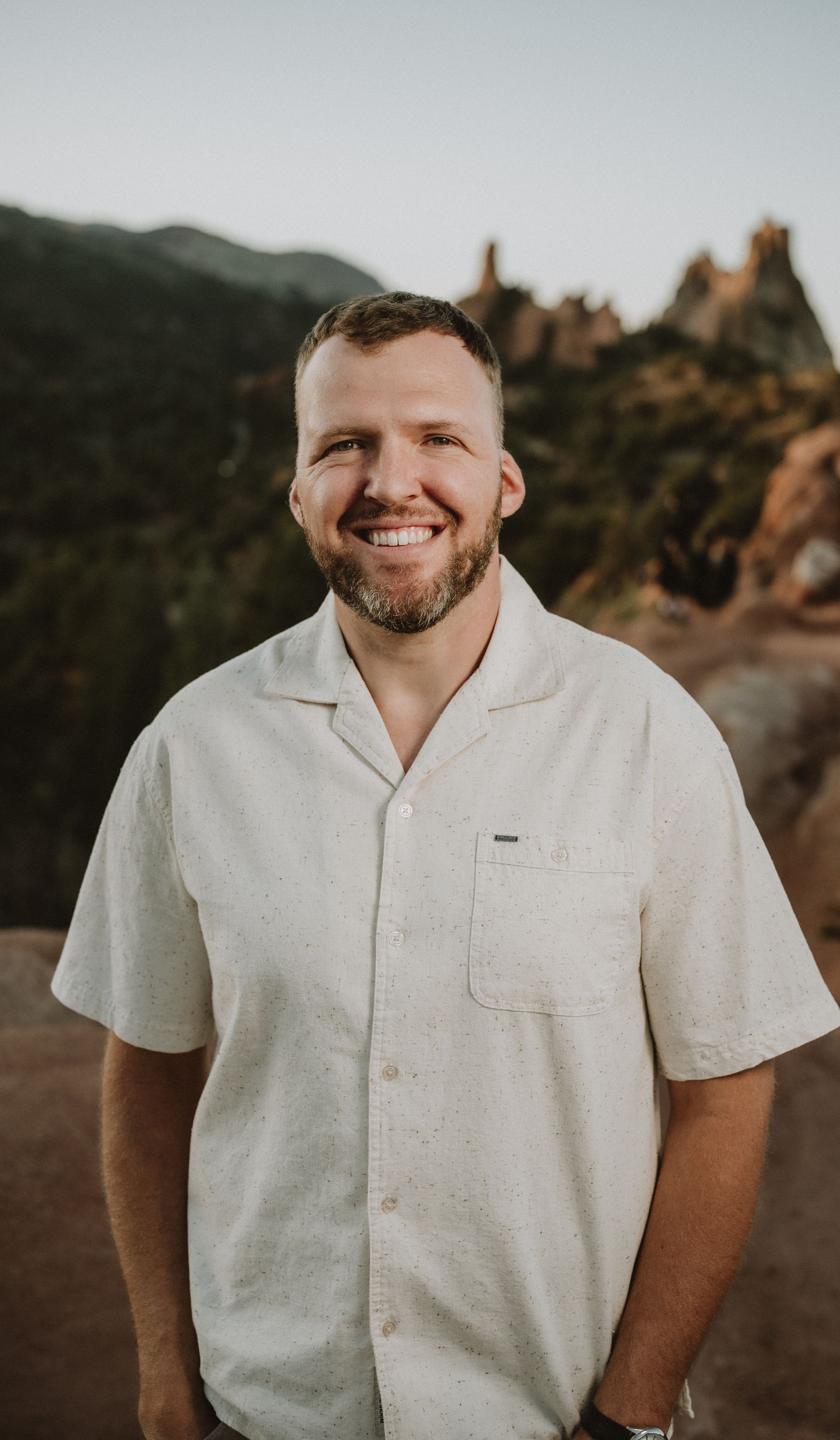 Man with a beard smiling outdoors, wearing a button-down shirt; background of red rocks.