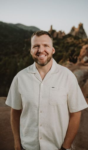 Man smiling, standing outdoors in front of a mountain, wearing a cream-colored button-up shirt.