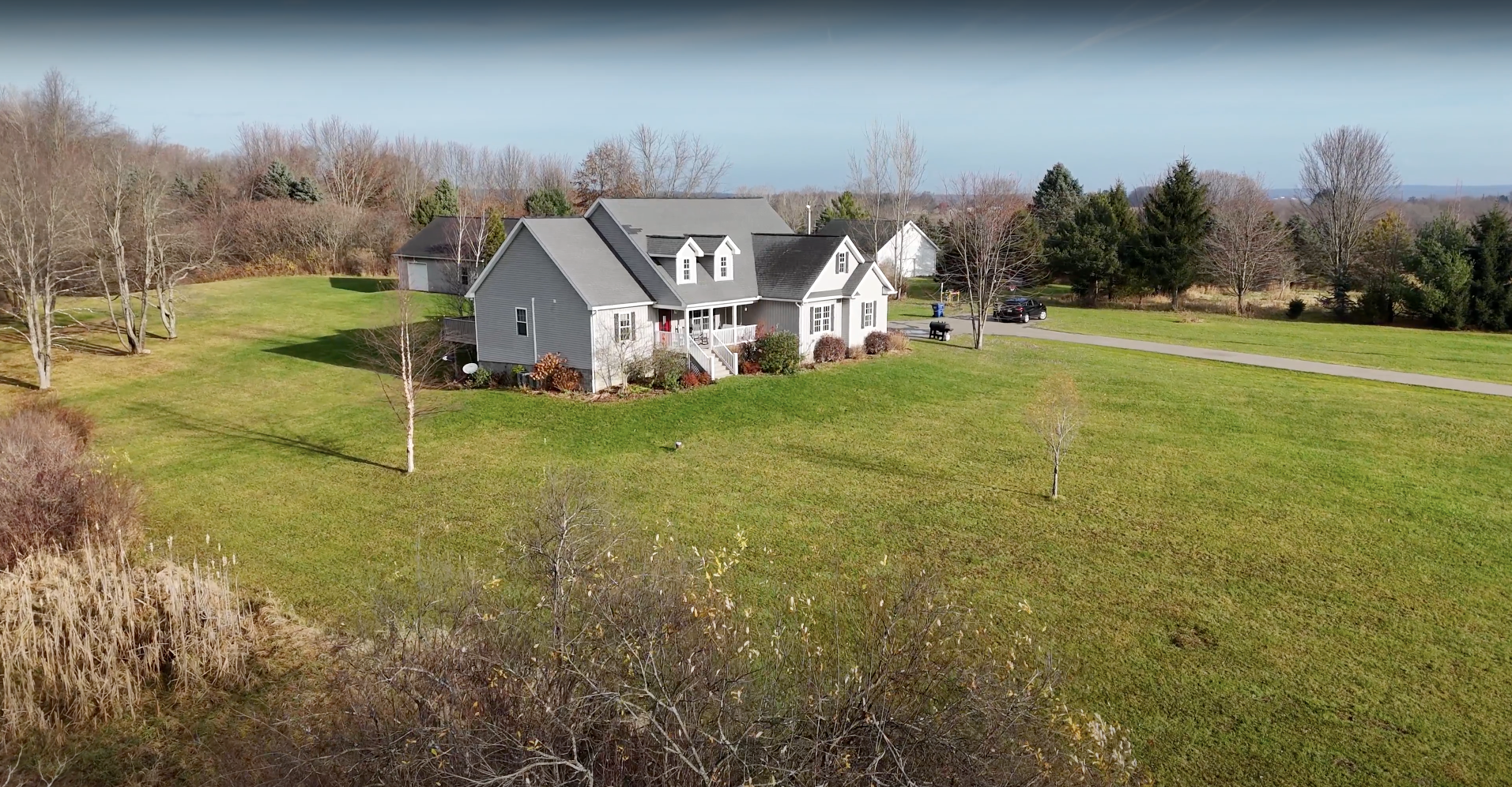 A gray house with a porch sits in a grassy field with a long driveway under a blue sky.