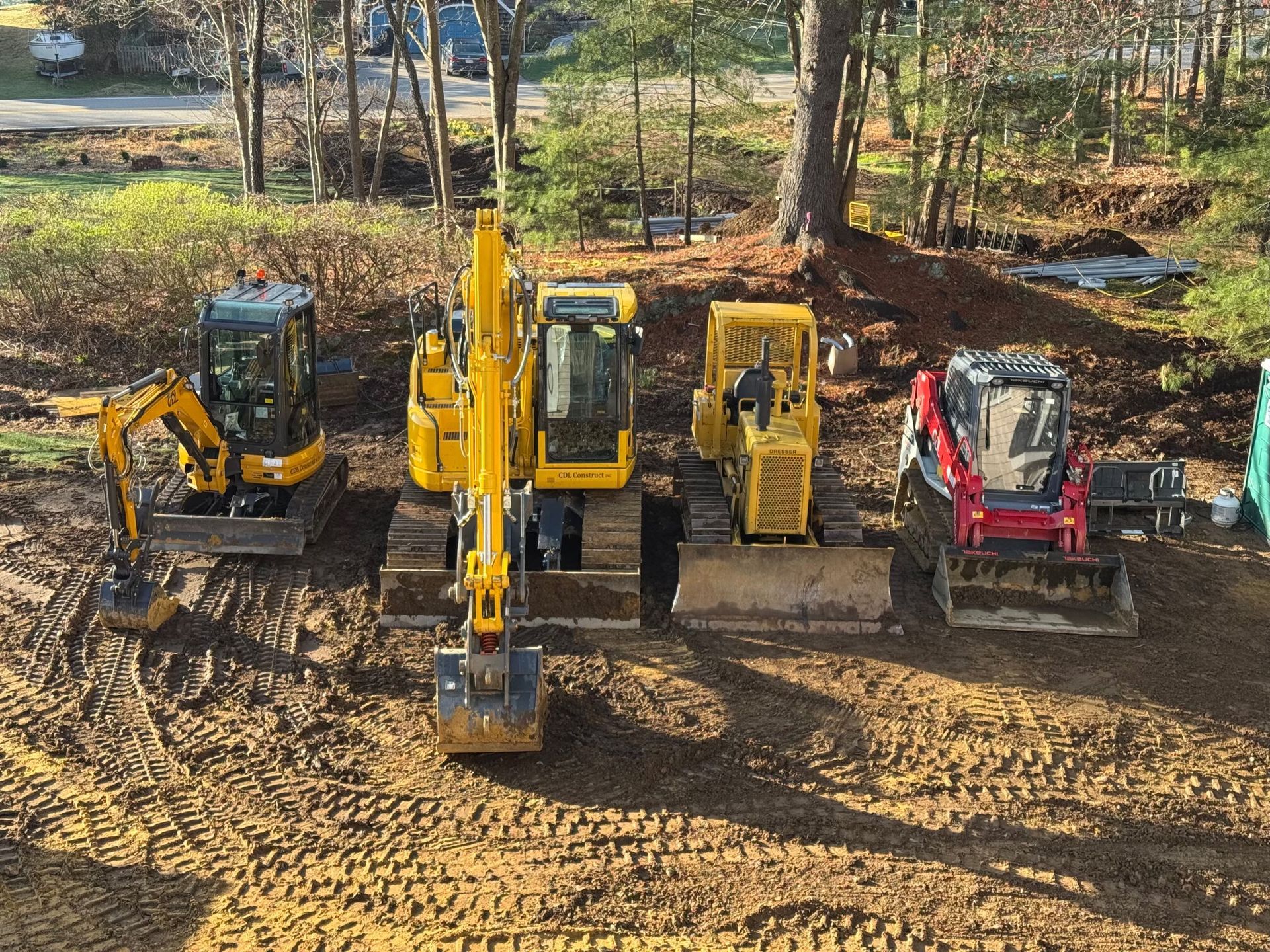 Four construction excavators and a skid steer parked on a dirt lot in a wooded area