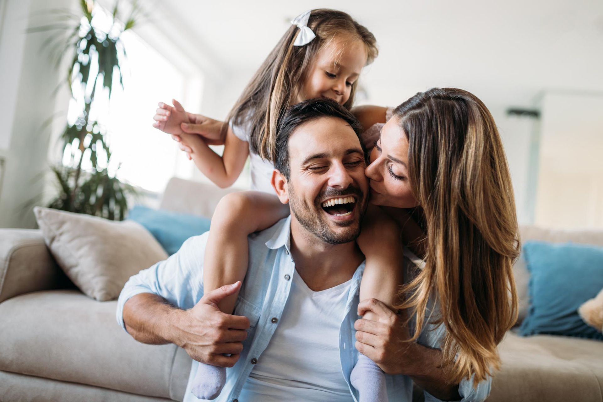 Smiling family on a couch, with a child on a man’s shoulders and a woman kissing his cheek
