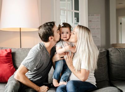 Family sitting on a couch, parents kissing a smiling child between them, in a bright living room