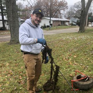 Man in work clothes holding debris, smiling, next to a drain-clearing tool in a grassy yard.