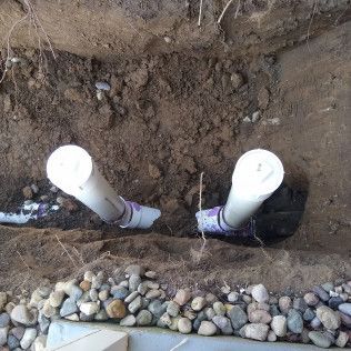 Two white PVC pipes emerge from the soil in a trench with gravel below.
