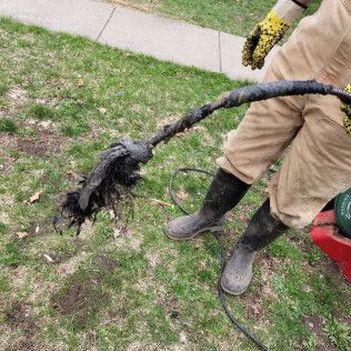 Person in work clothes holding a clogged drain snake with debris on a grassy lawn.