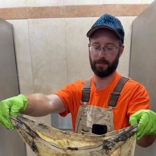 Man holding soiled cloth in bathroom, wearing gloves, orange shirt, overalls, and baseball cap.