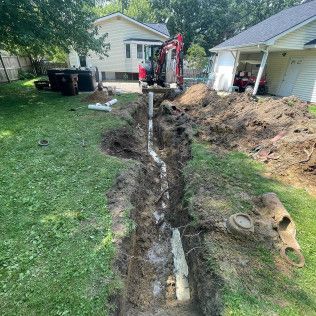 A backhoe digging a trench in a backyard, with pipes and dirt visible. Two houses are in the background.