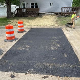 Asphalt patch on a street, with an orange traffic cone, and a worker using a power tool.