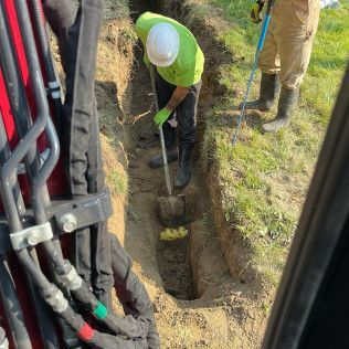 Two workers digging in a trench, one using a shovel, the other with a tool. Green grass surrounds them.