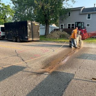 Man cutting asphalt with a saw, trailer and excavator in background.
