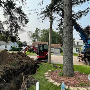 Mini excavator digs a trench in a grassy yard, near a tree. A pile of dirt is alongside. 