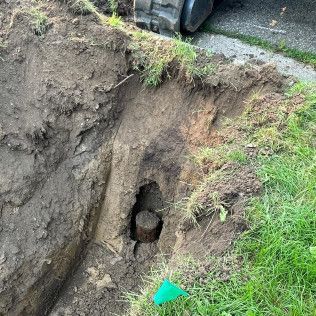 Hole in the ground revealing a cylindrical object, near grass and a sidewalk.